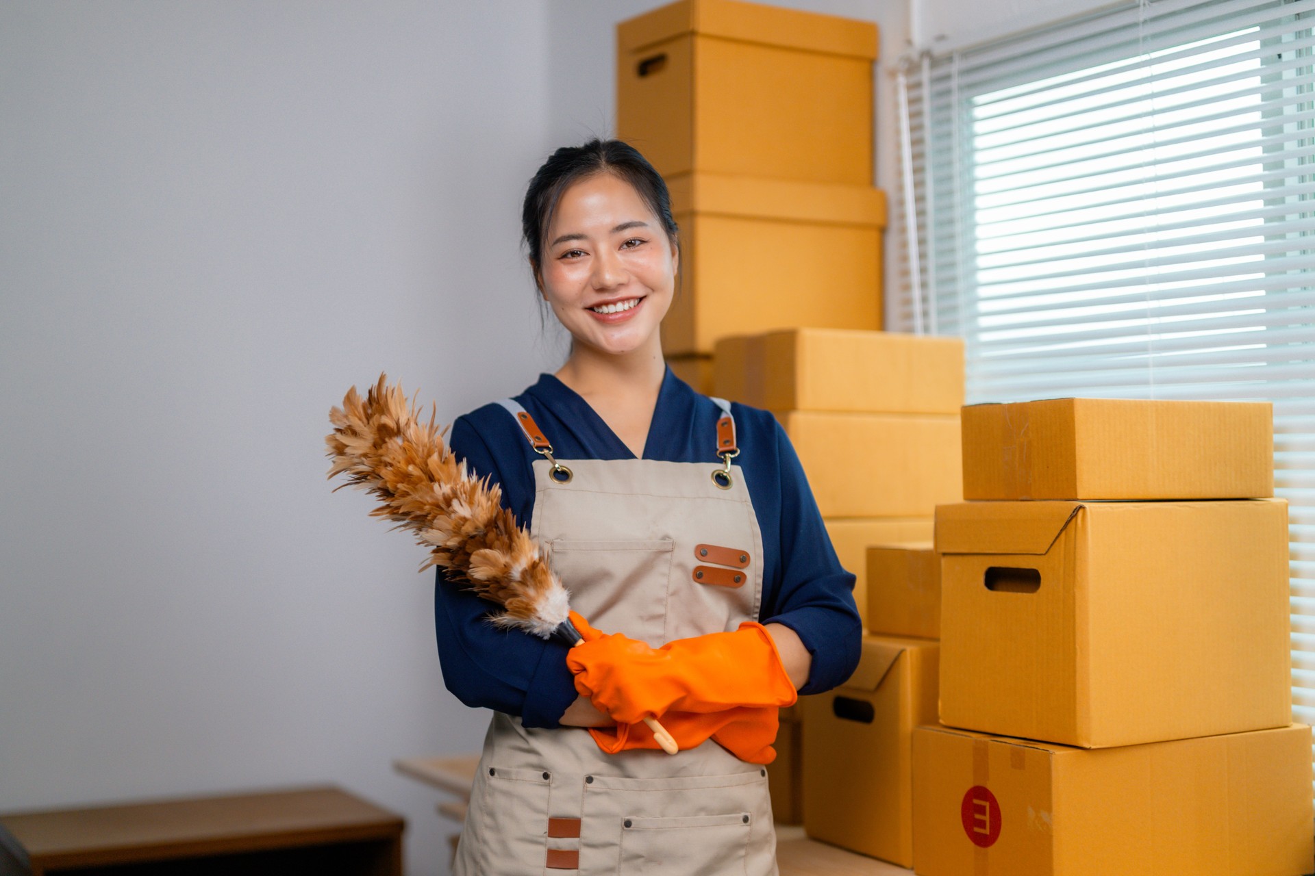 Smiling woman in a home office holding a duster, surrounded by cardboard boxes, showcasing cleaning and organizing